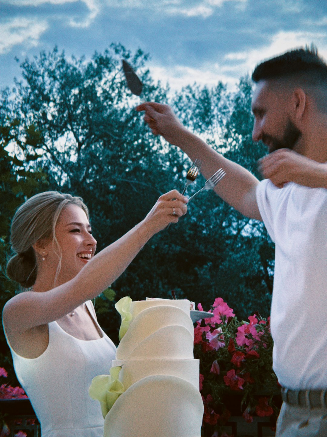 A woman and a man holding forks over a white cake outdoors