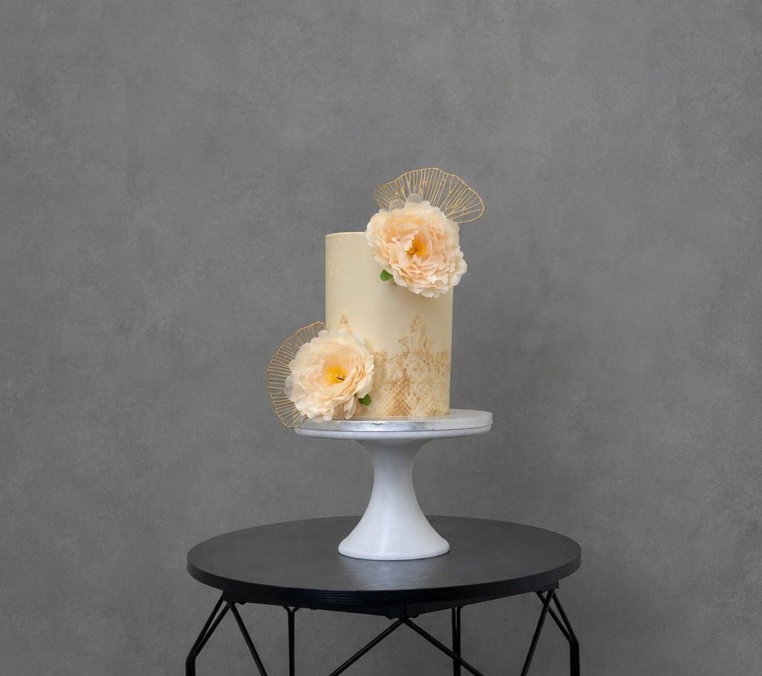 Tall ivory cake with two peach sugar flowers and gold lace fans on a white cake stand